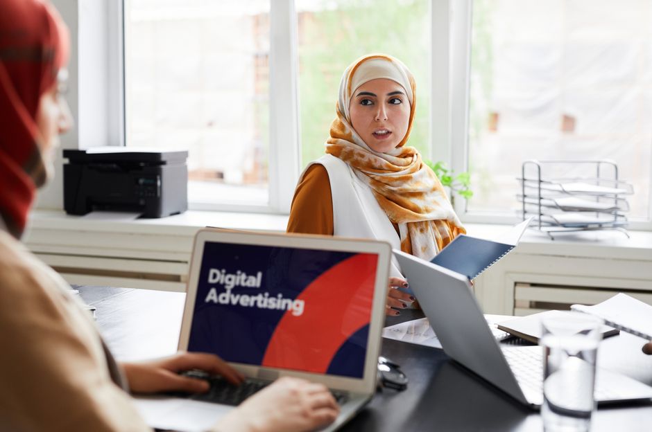 ai ad creative — Two women in hijabs discuss digital advertising in an office setting.
