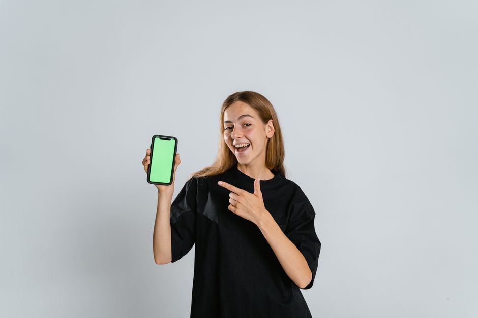 line lap audience — Smiling woman in black t-shirt pointing at a green screen smartphone indoors.