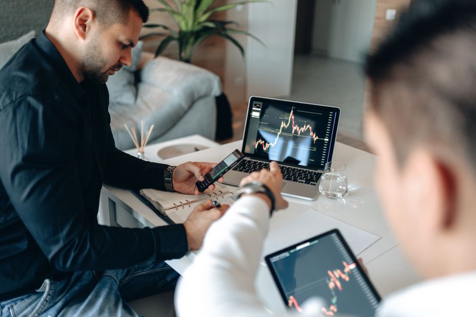 predictive analytics — Two businessmen analyzing financial data on laptops in a modern office setting.