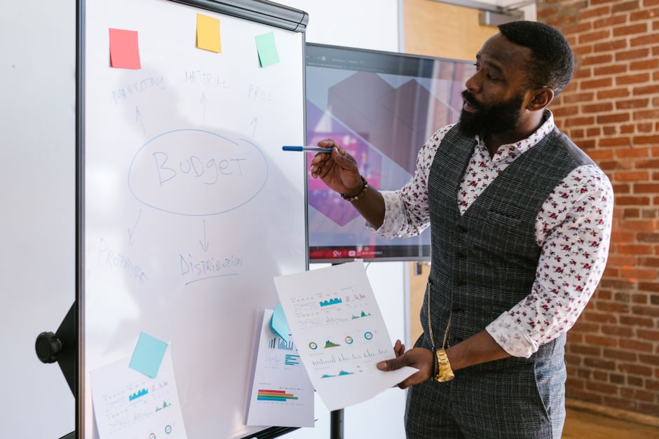 วางแผนงบการตลาด — A businessman explains budget strategies using a whiteboard in an office setting.
