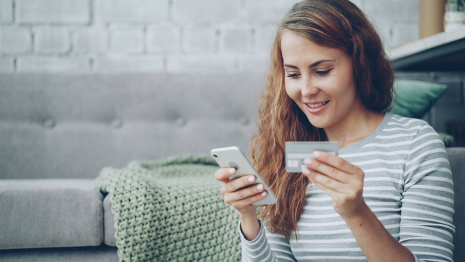 facebook shop ตั้งค่า — A smiling woman uses her smartphone and credit card for online shopping in a cozy living room.
