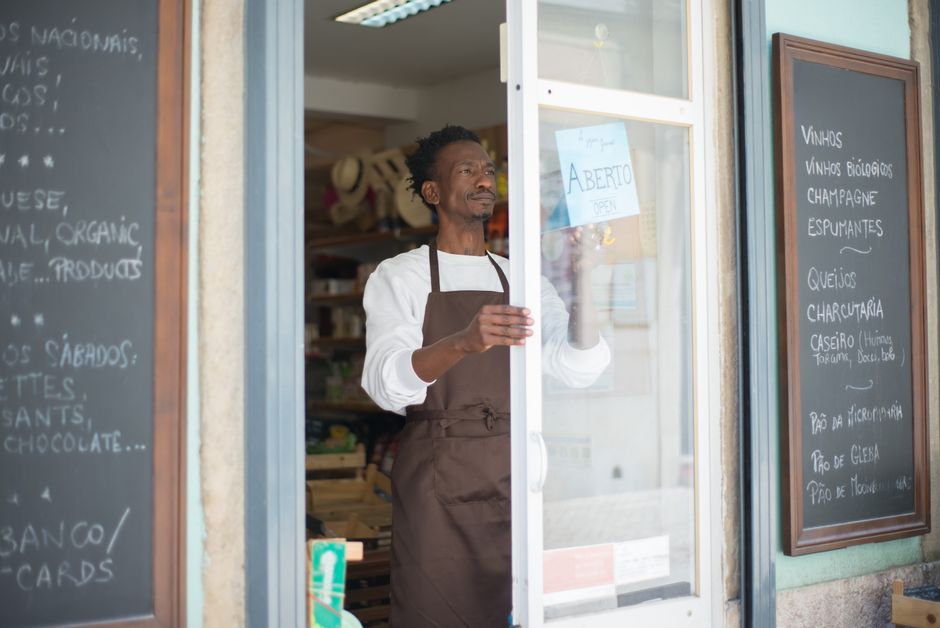 google business profile — A man in an apron opens a grocery store door in Portugal, ready for business.