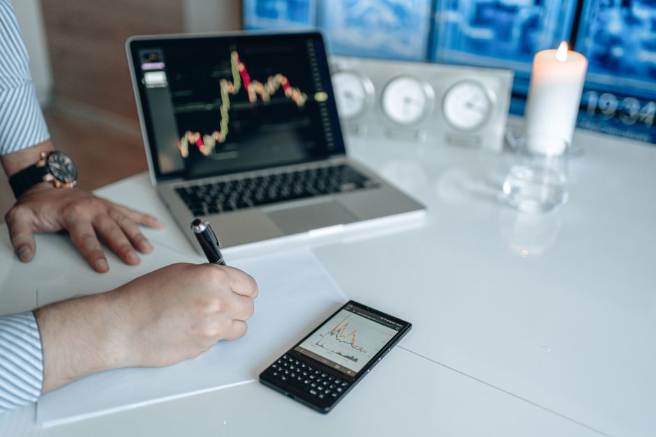 ltv คำนวณ — Business analyst writing with laptop and smartphone showing trend graphs on a white desk.