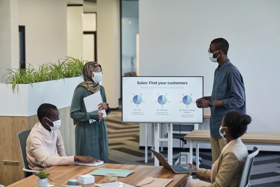 churn rate คือ — Business professionals in masks discuss customer profiles in a modern office setting.
