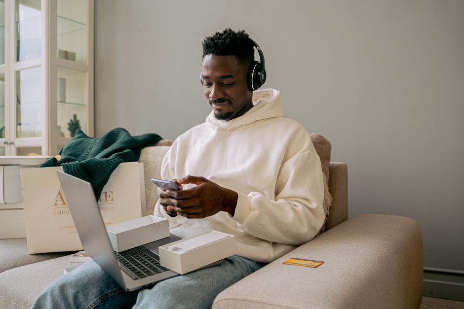 facebook shop ตั้งค่า — Man in a hoodie using a new smartphone and laptop on a sofa, indoors.